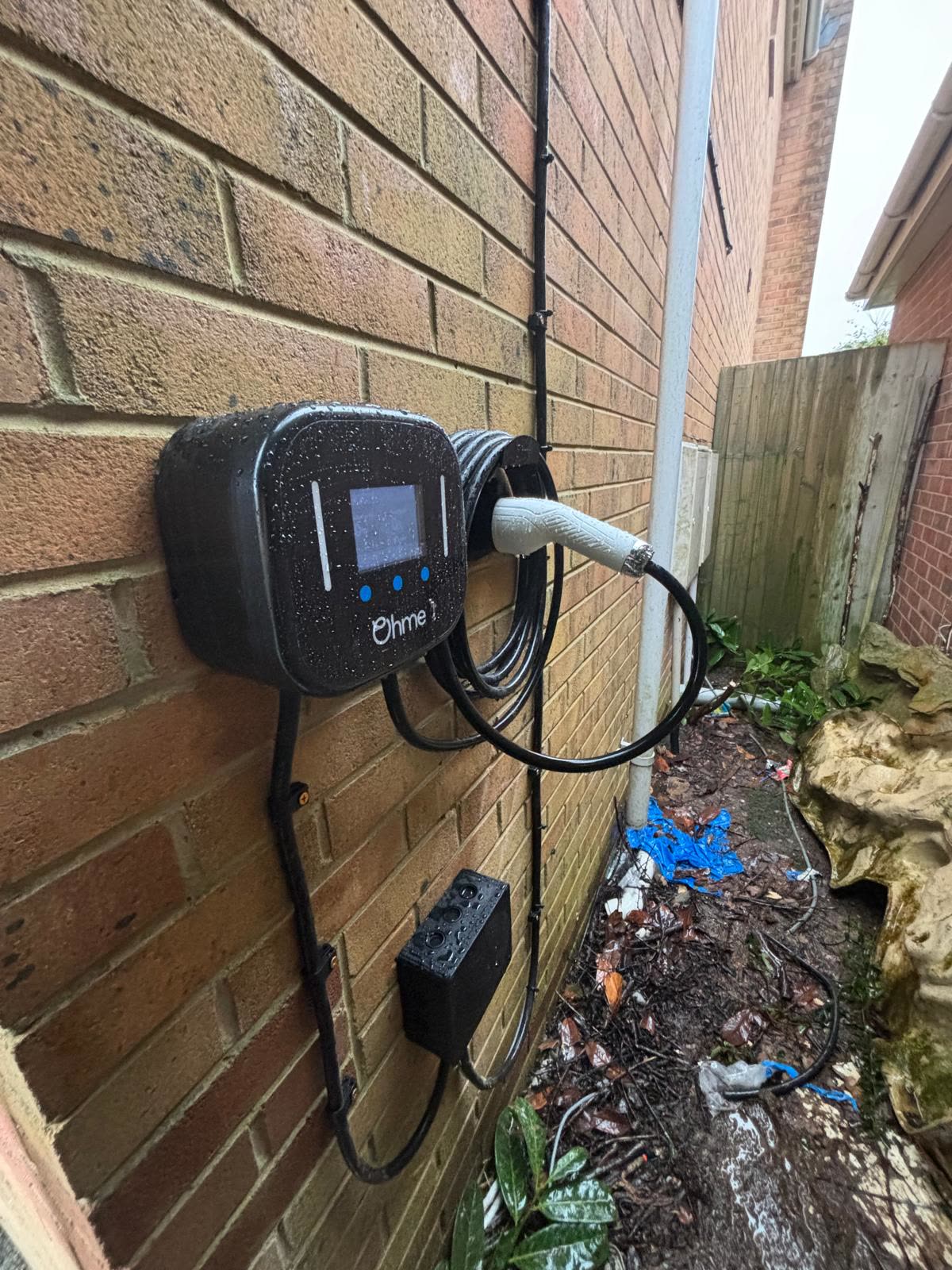 An Ohme EV charger fitted to a house wall by RM Electrical, cable coiled, on a wet day